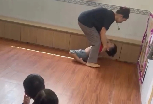 Bé trai bị cô giáo đè ra sàn, ngồi lên bụng, dùng đầu gối kẹp chặt đầu để nhét thức ăn vào miệng trong lúc bé đang khóc