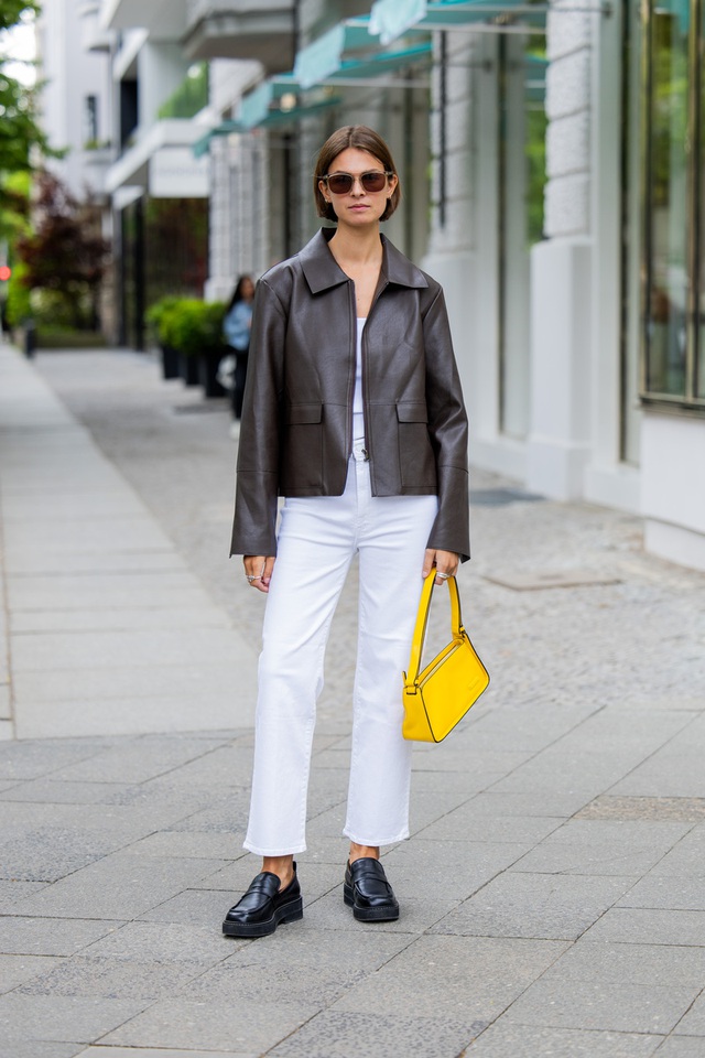 Một chiếc áo khoác da màu nâu kết hợp với quần jeans trắng để tạo nên sự tương phản rõ rệt, được tạo điểm nhấn bởi chiếc túi màu vàng mang lại màu sắc nổi bật